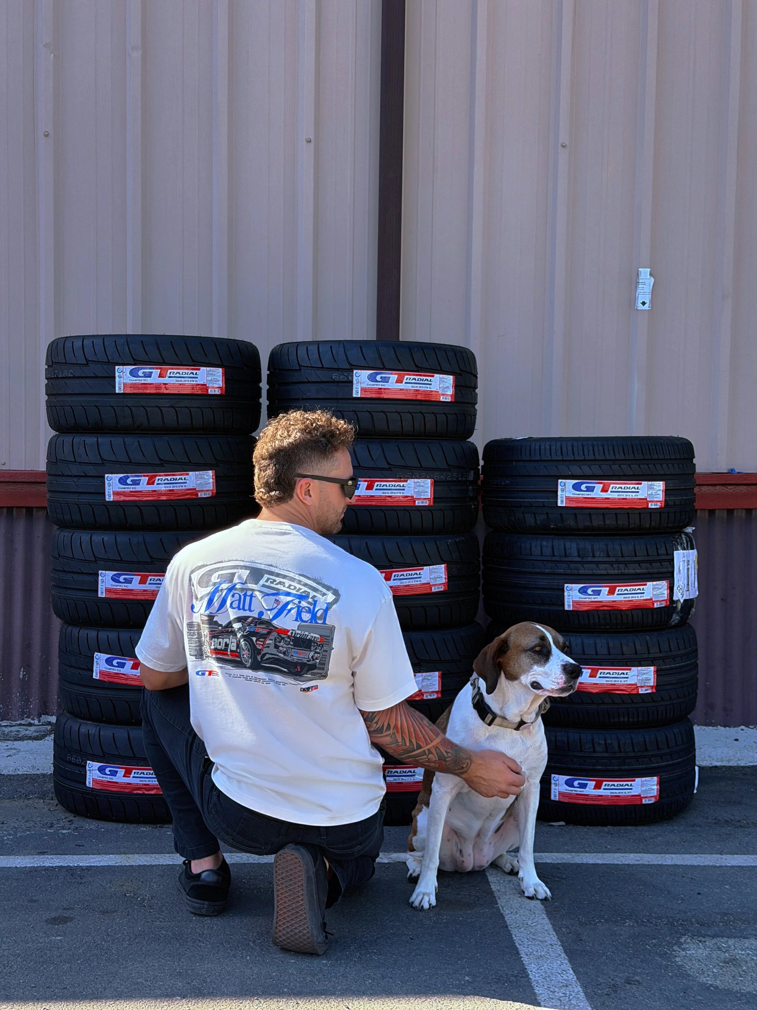 White tee with colorful car graphic on back, worn by person kneeling with a dog
